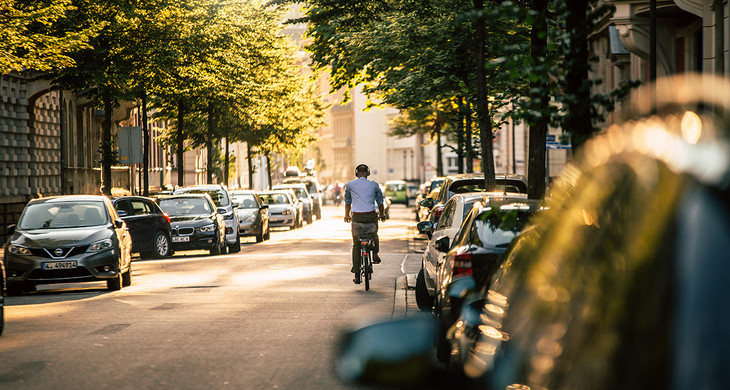 Fahrradfahrer mit Kopfhörern fährt auf einer Straße im Waldstraßenviertel. An den Seiten parken viele Autos.
