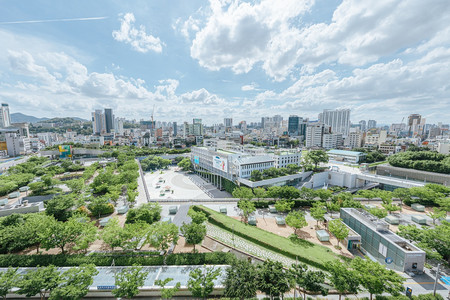 Blick auf ein großes modernes Gebäude des Asia Culture Center, das von einem Park umrundet ist. Im Hintergrund stehen weitere Wolkenkratzer der koreanischen Stadt Gwangju