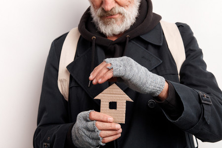 Ein älterer Mann, der einen Kapuzenpullover, Mantel und abgeschnittene Fingerhandschuhe trägt, hält eine Hand schützend über ein kleines Haus, ausgeschnitten aus Pappe, welches er in der anderen Hand hält.