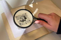 Eine Zeichnung in einem aufgeschlagenem Buch wird mit einer Lupe betrachtet