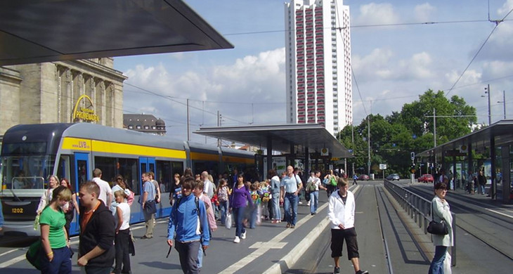 Menschen an der Straßenbahnhaltestelle am Leipziger Hauptbahnhof. Auf den Gleisen fährt eine Straßenbahn. Im Hintergrund ist das Wintergartenhochhaus.