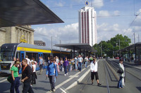 Menschen an der Straßenbahnhaltestelle am Leipziger Hauptbahnhof. Auf den Gleisen fährt eine Straßenbahn. Im Hintergrund ist das Wintergartenhochhaus.