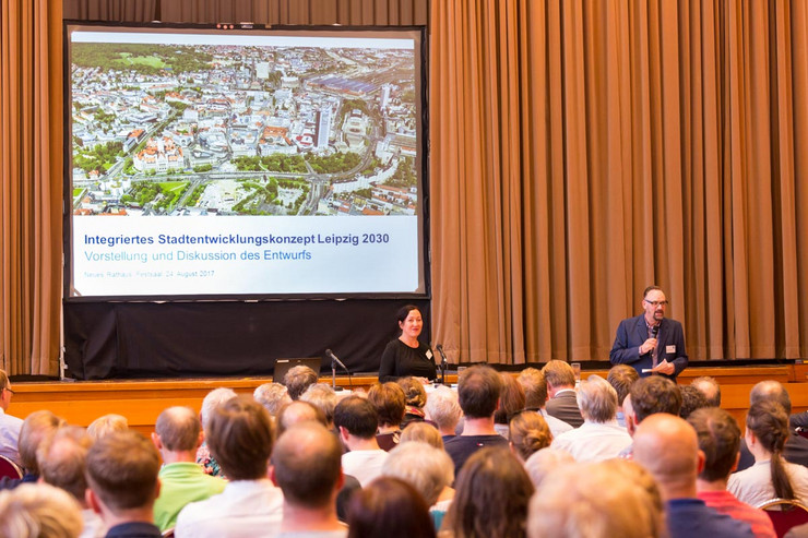 Eine Frau und ein Mann mit Mikrofon in der Hand stehen vor einem Publikum. Im Hintergrund eine Leinwand, auf der die Präsentation zum Stadtentwicklungskonzept präsentiert wird.