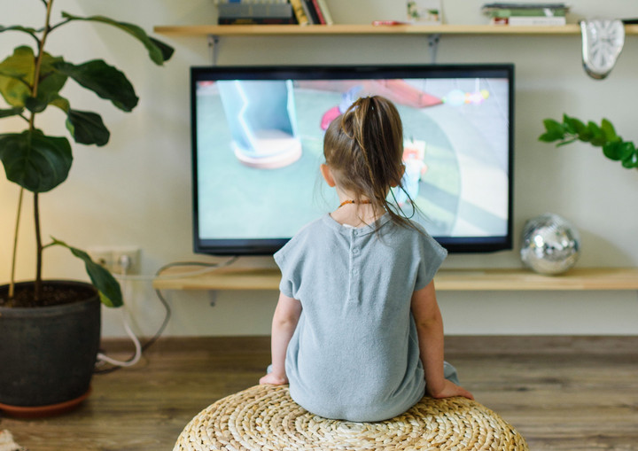 Ein Mädchen, das man von hinten sieht, sitzt vor einem Fernseher. Links ist eine grüne Topfpflanze. Der Fernseher ist an.