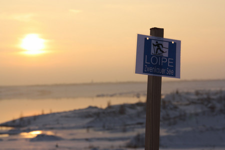 Hinweisschild für eine Ski-Langlauf-Loipe vor einer verschneiten Landschaft mit untergehender Sonne
