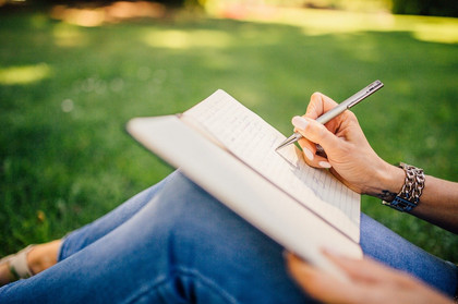 Bildausschnitt einer Person, die auf einer grünen Wiese sitzt und auf ein Blatt Papier schreibt