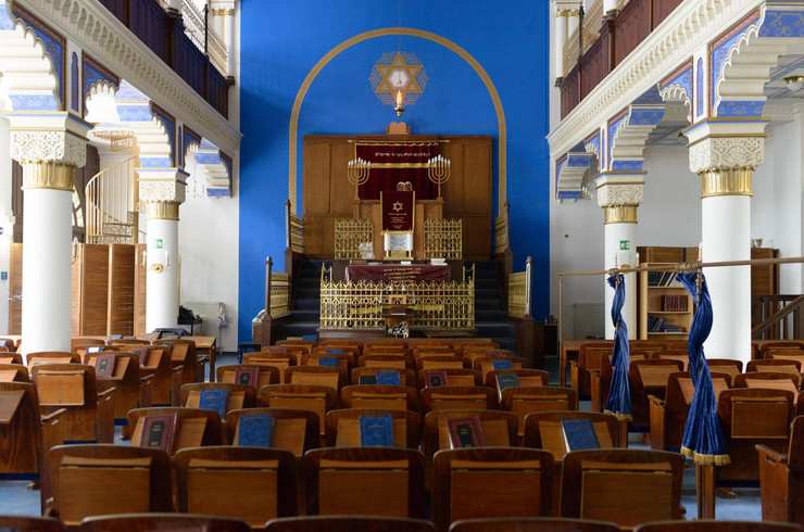 ein Synagogenraum mit Holzstühlen und Blick auf einen Thoraschrein vor blauer Wand mit Davidstern und siebenarmigen Kerzenleuchtern