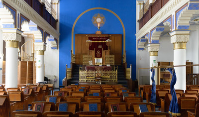 ein Synagogenraum mit Holzstühlen und Blick auf einen Thoraschrein vor blauer Wand mit Davidstern und siebenarmigen Kerzenleuchtern