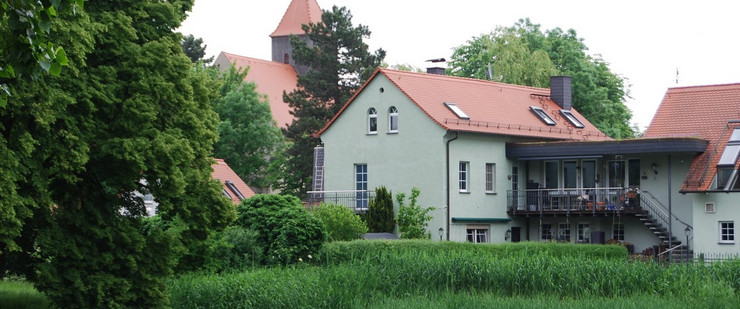 Ein Wohnhaus am Weiher in Holzhausen. Dahinter steht eine Kirche.