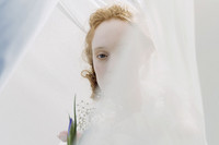 Eine blonde Frau mit einem Blumenstrauß hinter einem transparenten weißen Vorhang