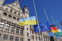 Die ukrainische Flagge in blau, gelb, die Europäische Flagge in blau und die Deutsche Flagge in Schwarz, Rot, Gold wehen auf halbmast vor einem Steingebäude mit mehreren Türmen. 