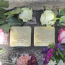 zwei goldene Steine mit der Inschrift in Erinnerung an die Verstorbenen, gesäumt von weißen und rosa Rosen