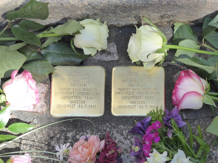 zwei goldene Steine mit der Inschrift in Erinnerung an die Verstorbenen, gesäumt von weißen und rosa Rosen