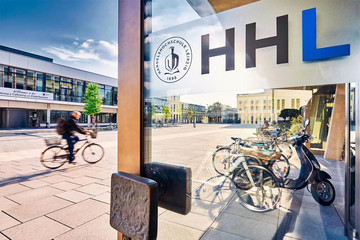 Ansicht des Campus' der HHL mit einem Mann auf einem Fahrrad und einer geöffneten Glastür mit dem Schriftzug "HHL"