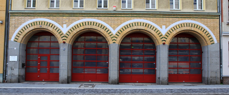 Das Gebäude der Freiwilligen Feuerwehr Ost mit vier großen, roten Rundbogentoren. Vor den Toren ist ein grau gepflasterter Fußweg.