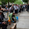 Ein Mann verlegt zwei Stolpersteine in den Boden eines Bürgersteigs. Einige Menschen stehen drum herum, eine Frau spielt Geige und ein Mann Ziehharmonika.