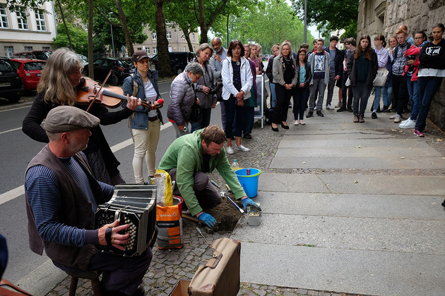 Ein Mann verlegt zwei Stolpersteine in den Boden eines Bürgersteigs. Einige Menschen stehen drum herum, eine Frau spielt Geige und ein Mann Ziehharmonika.