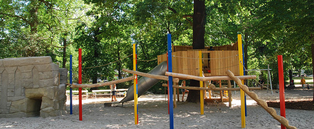 Klettermöglichkeiten und ein Baumhaus mit Rutsche unter einem großen Baum auf dem Spielplatz am Rennbahnweg