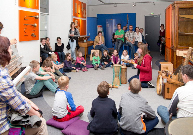 Kinder und ein paar Erwachsene sitzen im Kreis in einem Ausstellungsraum für Musikinstrumente. In der Mitte sitzt eine Frau, die verschiedene Instrumente zeigt und erklärt.