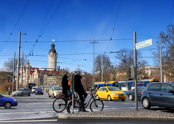 Blick auf eine große Straßenkreuzung in Leipzig mit Autos, Radfahrern und Straßenbahnen. Im Hintergrund das Neue Rathaus.