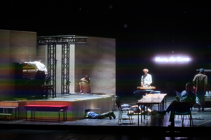 Theaterstück auf einer Bühne: zwei Menschen liegen am Boden, einer sitzt am Tisch und liest Zweitung, einer steht am Klavier.