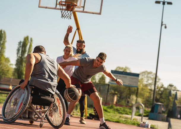 Ein Mann im Rollstuhl spielt mit drei Personen im Freien Basketball. Eine Mitspielerin lächelt.
