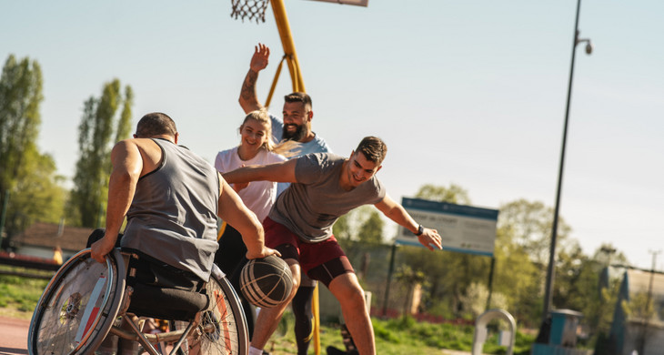 Ein Mann im Rollstuhl spielt mit drei Personen im Freien Basketball. Eine Mitspielerin lächelt.