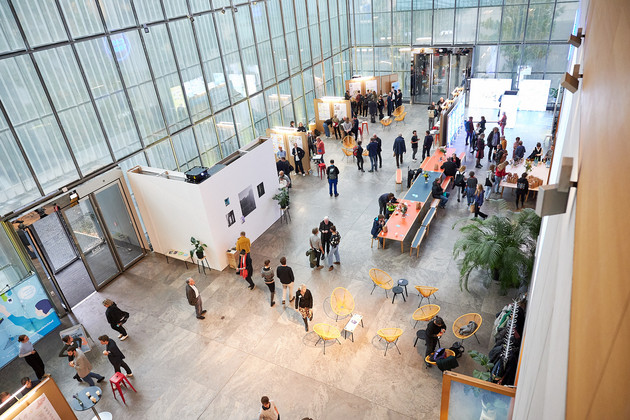 Blick von oben in eine große Halle mit Ausstellungsflächen und Tischen und Menschen im Gespräch