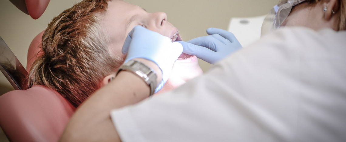 Ein Kind mit geöffnetem Mund auf einem Behandlungsstuhl beim Zahnarzt. Die Zahnärztin beugt sich über das Kind und schaut in seinen Mund.