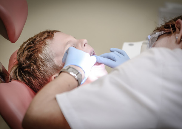 Ein Kind mit geöffnetem Mund auf einem Behandlungsstuhl beim Zahnarzt. Die Zahnärztin beugt sich über das Kind und schaut in seinen Mund.