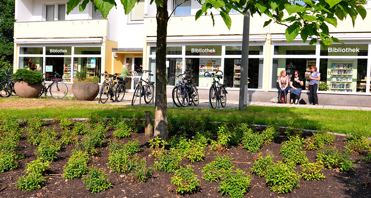 Blick über eine gepflegte Grünanlage auf die Fensterfront der Bibliothek Schönefeld im Erdgeschoss eines Plattenbaus. Vor der Bibliothek eine Gruppe von drei Personen und Fahrradständer.