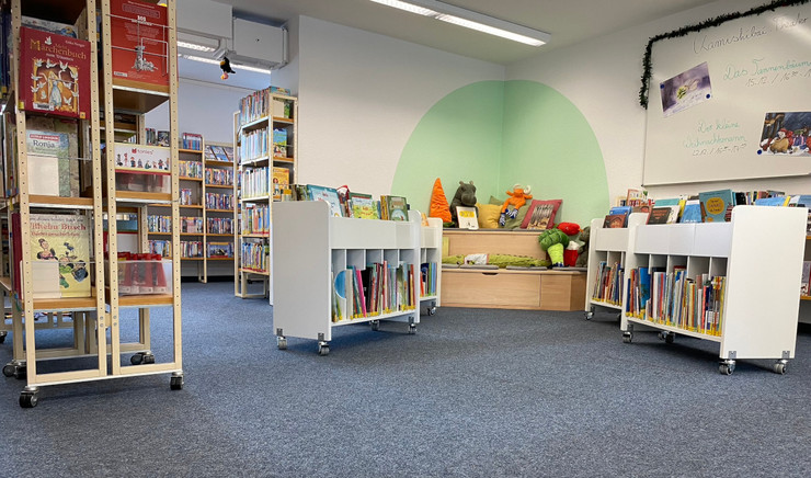 Kinderbibliothek mit Leseecke, großen Bücherregalen, kleinen Rollregalen und einer großen Tafel.