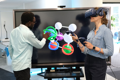 Mann in weißem Hemd und Frau mit 3D-Brille und Eingabegeräten vor großem Computerbildschirm, der eine 3D-Grafik aus bunten. molekülartig verbundenen Kugeln zeigt.