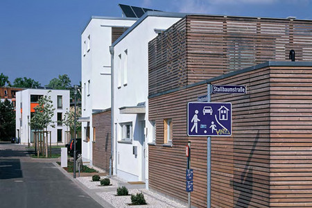 Blick in die Stallbaumstraße: vorne ein Spielstraßenschild, rechts ein Stadthaus aus Holz, dahinter weiße Stadthäuser