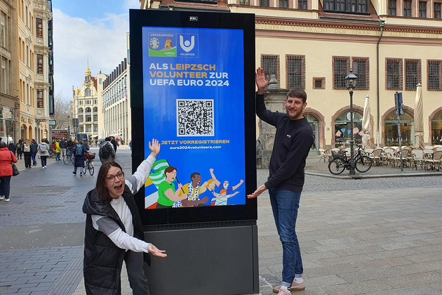Zwei Personen präsentieren ein Werbeplakat zum Thema Volunteer Vorregistrierung in der Leipziger Innenstadt