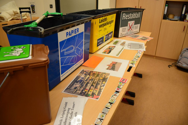 Auf einem Tisch sind mehrere verschiedene Abfallbehälter aufgereiht wie für Plastik, Biomüll und Papier. Davor liegen kleine und große Bildkarten.
