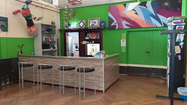 In einem Raum mit grünen Wänden steht eine Bar mit vier Hockern. An der linken Wand hängt eine Pappmascheefigur. Eine offene Tür führt in eine Küche.