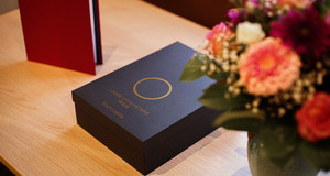 Ein Blumenstrauß, der Preisgegenstand und die Urkunde in rotem Einband auf einem Tisch