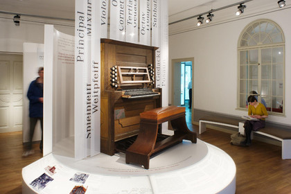auf einer Empore steht ein historisches Klavier, dahinter beschriftete Tafeln