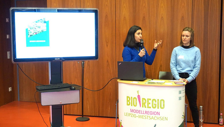 Zwei Mitarbeiterinnen der Bio-Regio-Modellregion Westsachsen stehen an ihrem Infostand. Eine von Ihnen spricht in ein Mikrofon. Links neben Ihr steht ein Präsentationsmonitor auf einem Rollgestell.