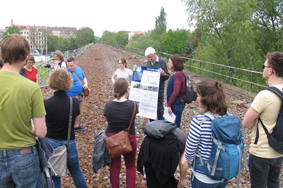 Eine Gruppe Menschen steht auf dem alten Gleisbett ehemaliger Schienen. Ein Mann erklärt etwas anhand eines Infoplakats, das er festhält.