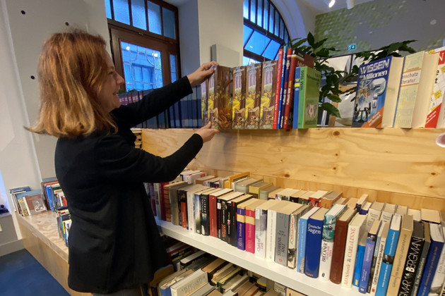 Eine Frau steht vor einen Bücherregal und greift nach einem Buch.