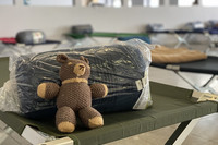Auf einem Feldbett liegt ein verpackter Schlafsack und ein Teddybär
