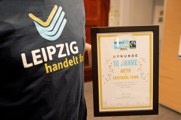 Die gerahmte Urkunde "10 Jahre Fairtrade-Town Leipzig" in der Nahaufnahme, daneben ist ein T-Shirt mit dem Aufdruck "Leipzig handelt fair" zu erkennen.