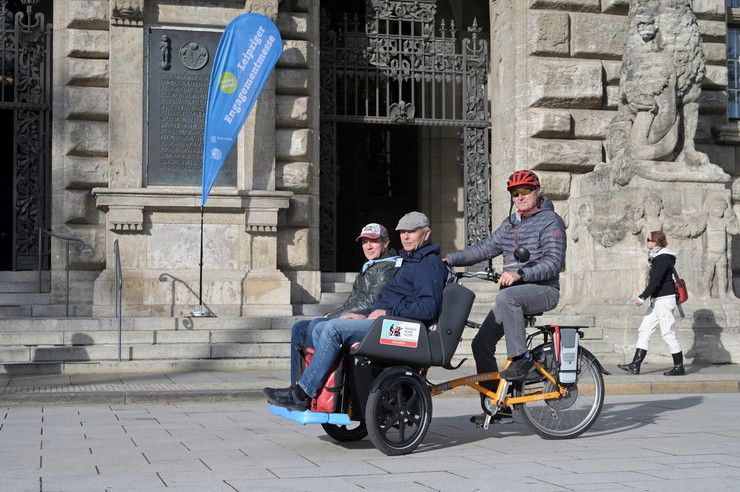 Zwei Personen werden von einer dritten Person mit einer Rikscha vor dem Neuen Rathaus zu Leipzig transportiert.
