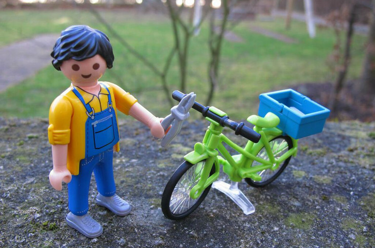 Playmobilfigur mit gelbem Oberteil und Blaumann repariert ein grünes Fahrrad.