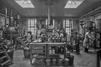 Schwarz-Weiß-Foto. Blick in die Fabrik für ätherische Öle in der Mahlmannstraße im Jahr 1920. Ein Raum mit vielen Kesseln, Glasflaschen und Metallbehältern, in dem Menschen in weißen Kitteln arbeiten.