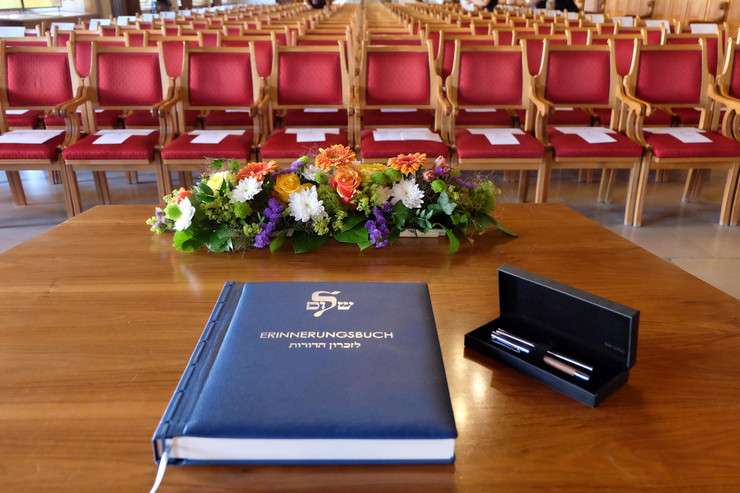 blaues Buch mit goldener Schrift, davor Blumen auf einem braunen Tisch und im Hintergrund zahlreiche leere, rote Stühle zu sehen