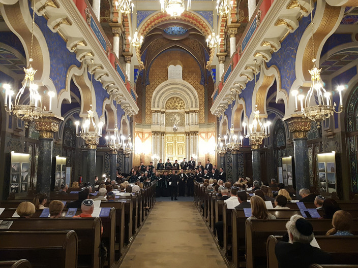 Blick inmitten der dunklen, vollbesetzten Sitzreihen und blau-goldenen Wände auf den Chor, der in schwarzen Gewändern singt