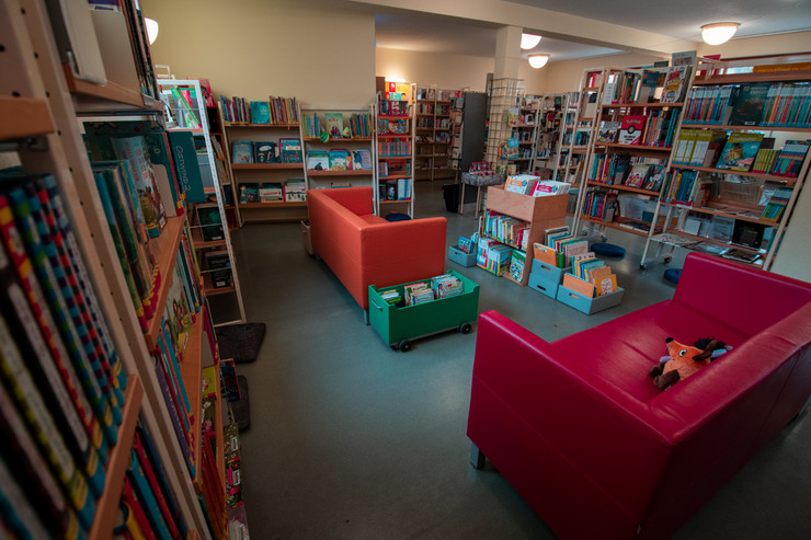 Blick in die Kinderbibliothek. Mehrere Regale mit Büchern umrahmen den Innenbereich mit zwei roten Couches.